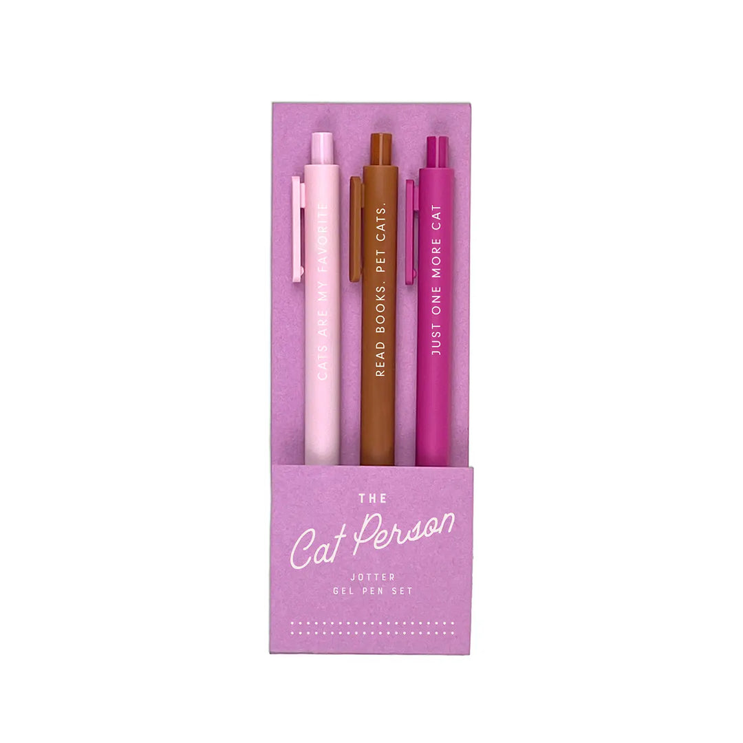 A set of three gel pens in a pastel purple packaging labeled "The Cat Person Jotter Gel Pen Set." The pens come in soft pink, warm brown, and deep magenta, each featuring a playful phrase in white lettering: "Cats Are My Favorite," "Read Books. Pet Cats.," and "Just One More Cat."