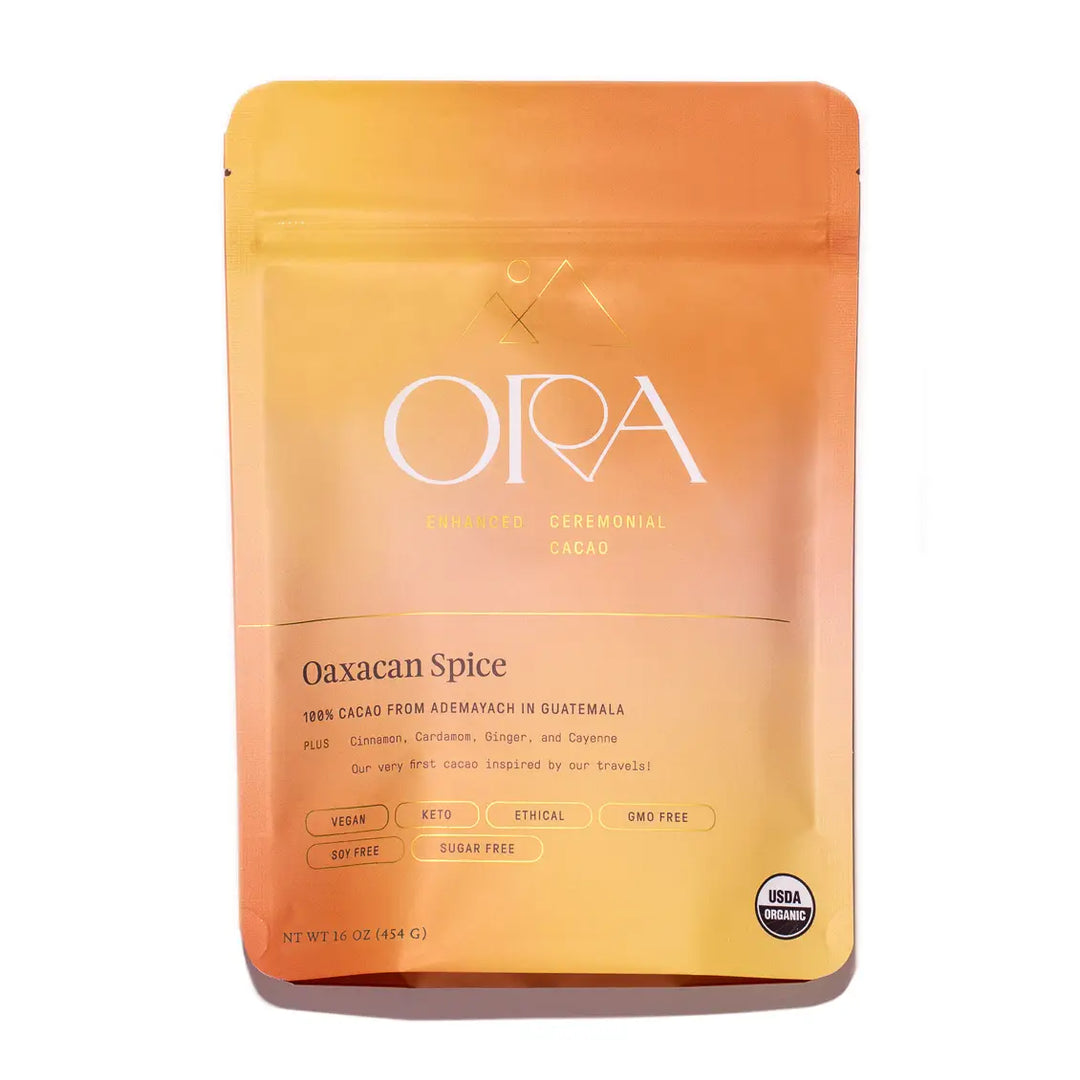 A vibrant orange and yellow resealable pouch labeled "ORA Enhanced Ceremonial Cacao, Oaxacan Spice." The packaging highlights its 100% cacao sourced from Ademayach in Guatemala, with added spices like cinnamon, cardamom, ginger, and cayenne. USDA Organic, vegan, keto-friendly, and free of sugar, soy, and GMOs.