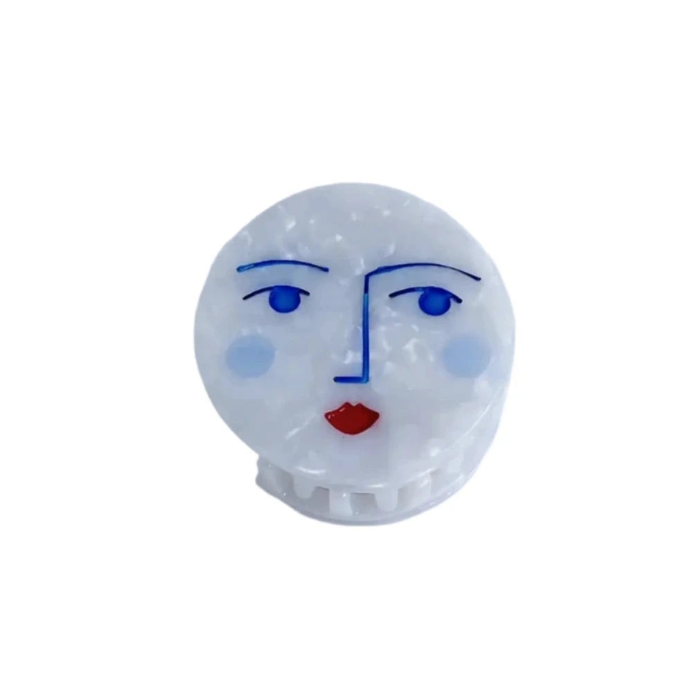 A hair claw with a playful moon face design featuring blue eyes, a red mouth, and minimalist features, set against a white background.