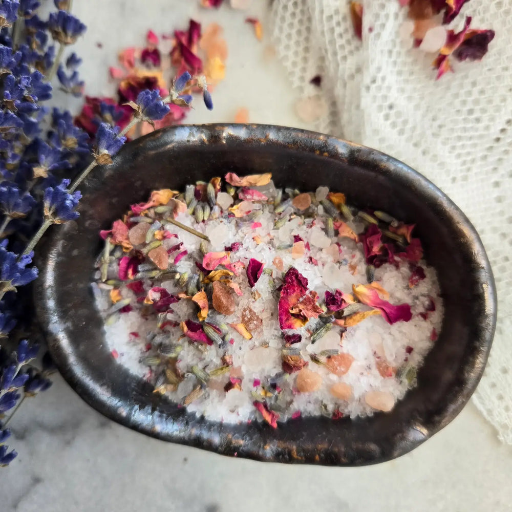 A close-up of a black ceramic dish filled with botanical bath soak, featuring pink Himalayan salt, dried rose petals, lavender buds, and other natural ingredients. Lavender sprigs and delicate lace fabric are in the background.