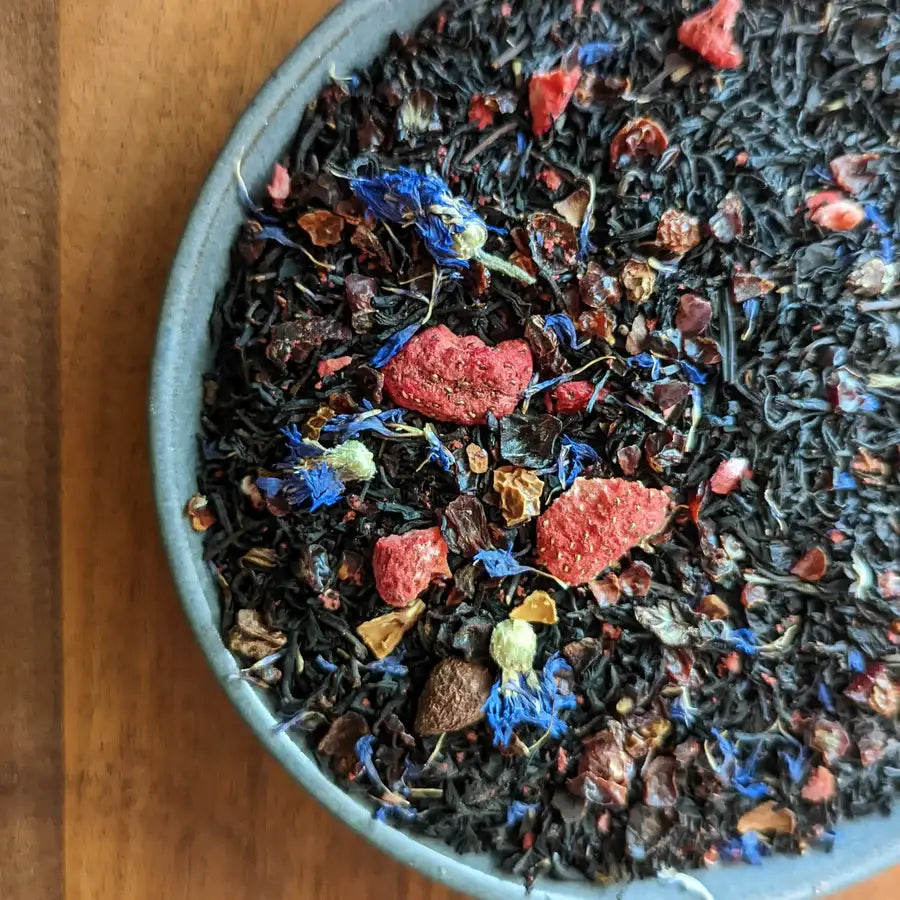 A close-up of a ceramic bowl filled with loose leaf black tea, featuring dried strawberries, blue cornflower petals, and other botanicals. The bowl is placed on a wooden surface.