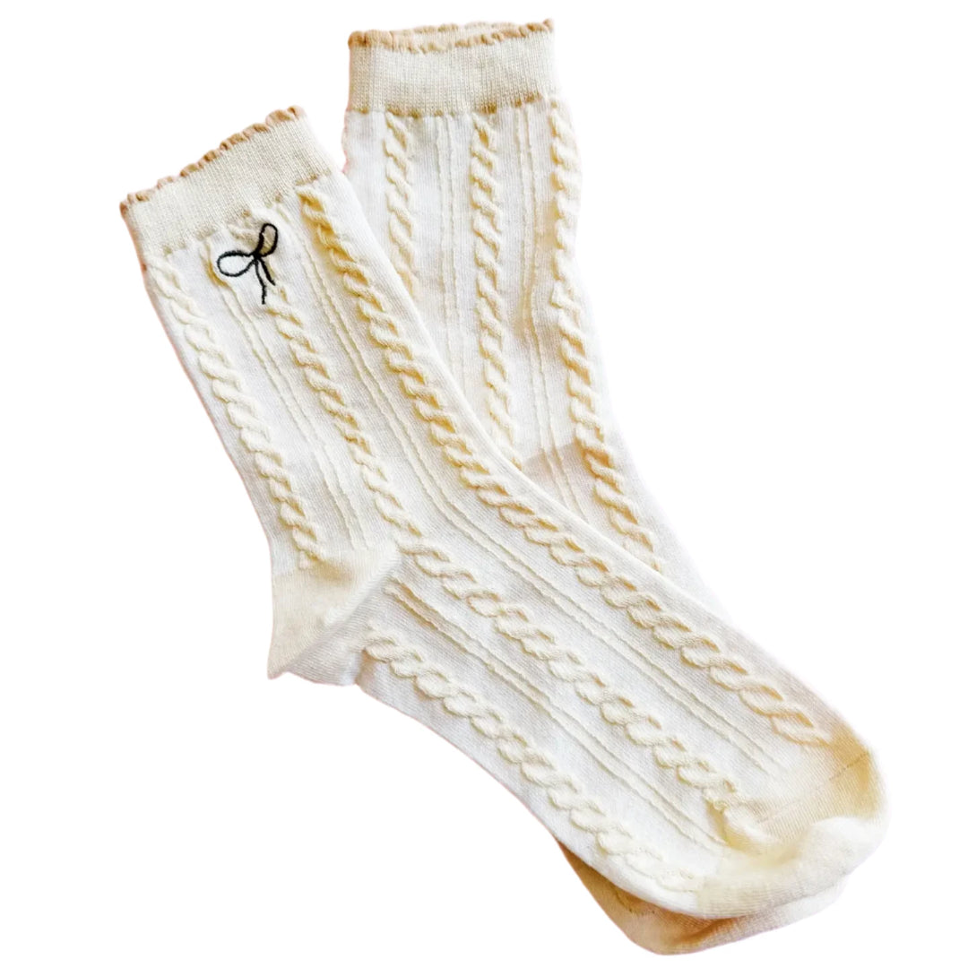 A pair of cream-colored cable-knit socks with delicate detailing, featuring a small embroidered black bow near the top. The socks are laid out on a soft pink background, showcasing their cozy and elegant design.