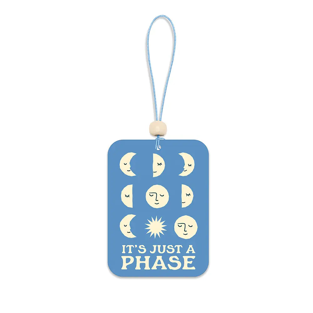Blue rectangular car air freshener with cream moon-phase illustrations, a small sun icon, and the phrase “It’s Just a Phase,” hanging from a light blue string with a wooden bead.