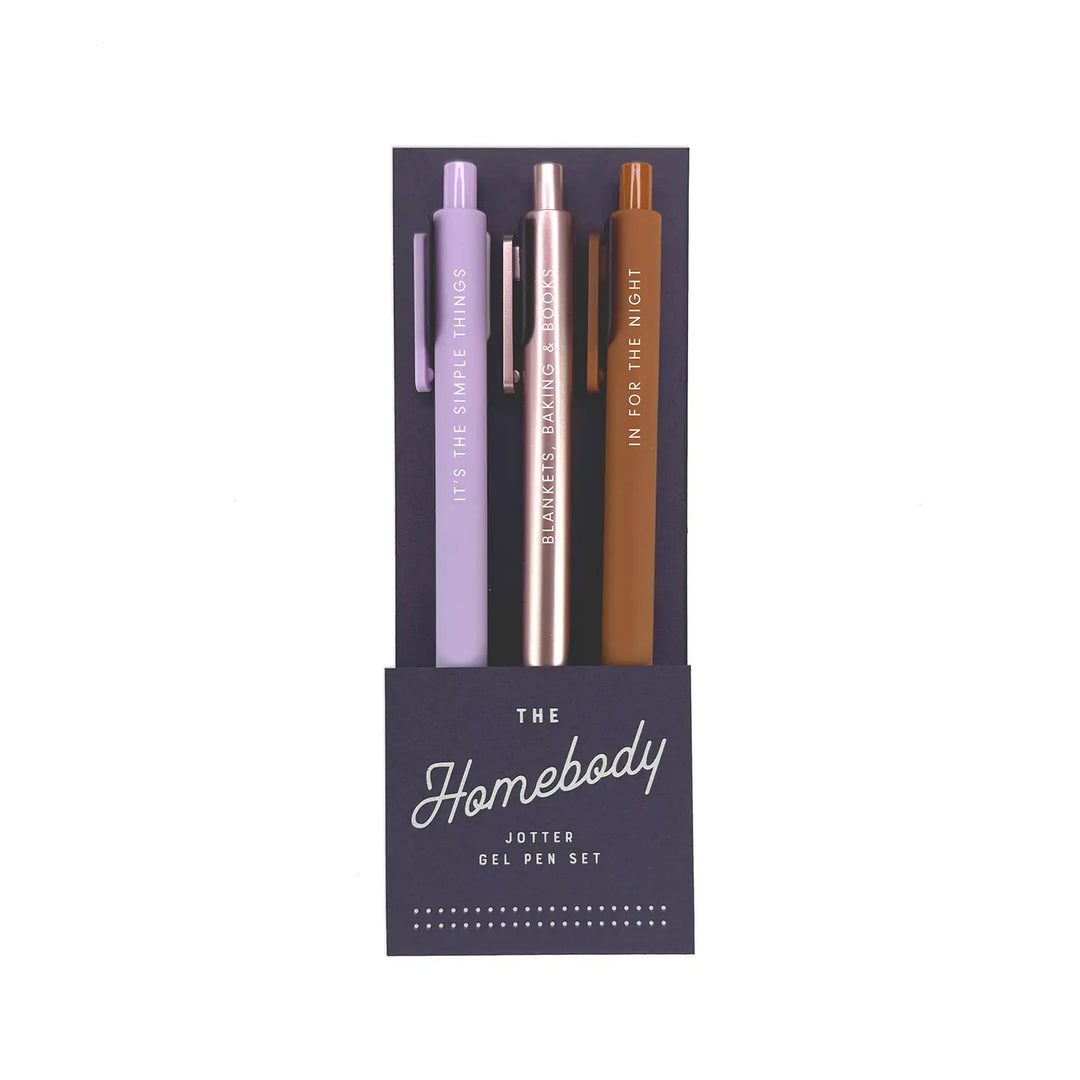 A set of three gel pens in a sleek, dark navy packaging labeled "The Homebody Jotter Gel Pen Set." The pens come in soft lavender, metallic rose gold, and warm brown, each featuring a cozy phrase in white lettering: "It's the Simple Things," "Blankets, Baking & Books," and "In for the Night."