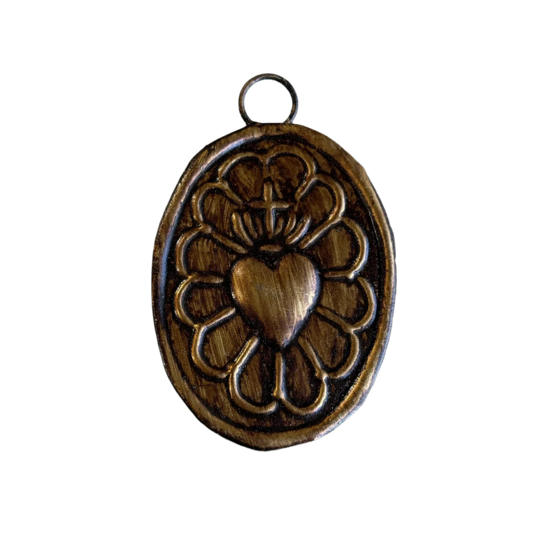 An oval-shaped bronze pendant featuring a raised design of a sacred heart surrounded by decorative floral patterns, with a small loop at the top for hanging or wearing.