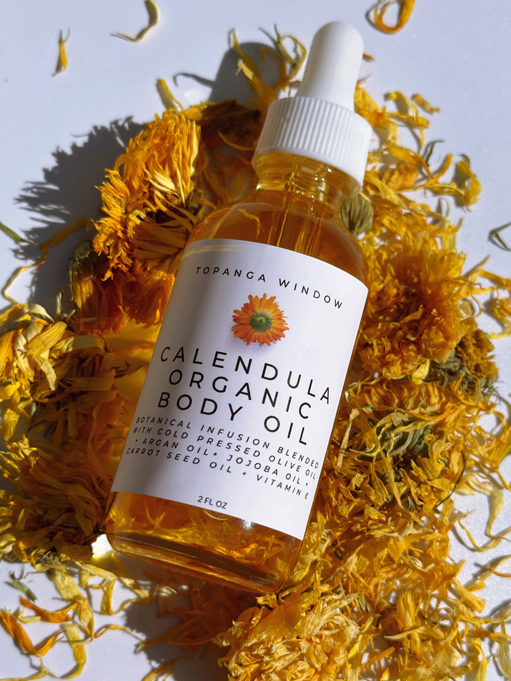 close up view of a 2 oz clear bottle of calendula organic body oil laying on a bed of dried calendula petals. Brand: Topanga Window