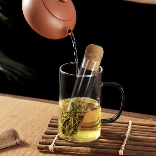 perforated glass tube tea infuser filled iwth loose leaf tea sitting in a glass mug with water being poured onto it.