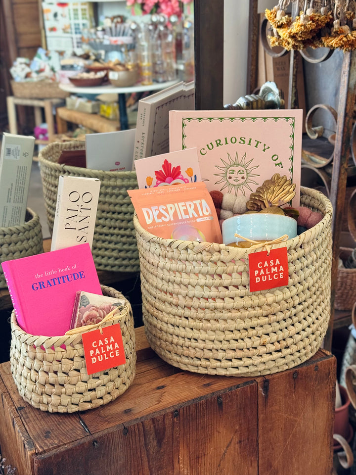 Two handwoven palm leaf baskets of different sizes styled as Casa Palma Dulce gift baskets filled with books, a Despierta superpowder blend, a candle, and assorted goods, displayed in a boutique setting.