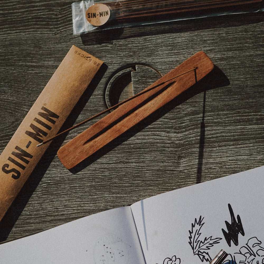 photo of a desk with a package of incense, an incense stick burning on a wooden incense holder and the corner of a sketchbook. Brand: Sin-min