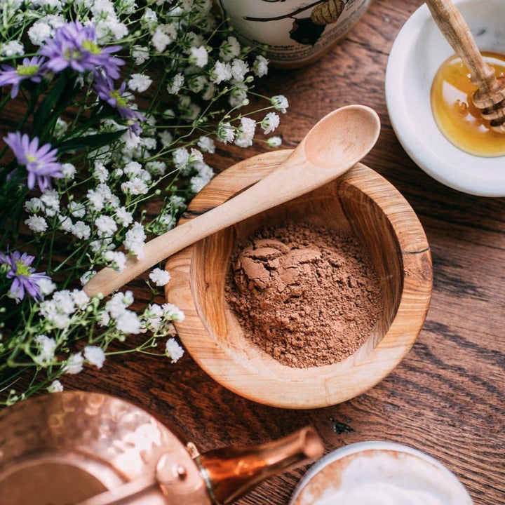 photo of a woooden bowl  and spoon filled with cacao powder and baby breath and purple flowers to the left of the bowl.