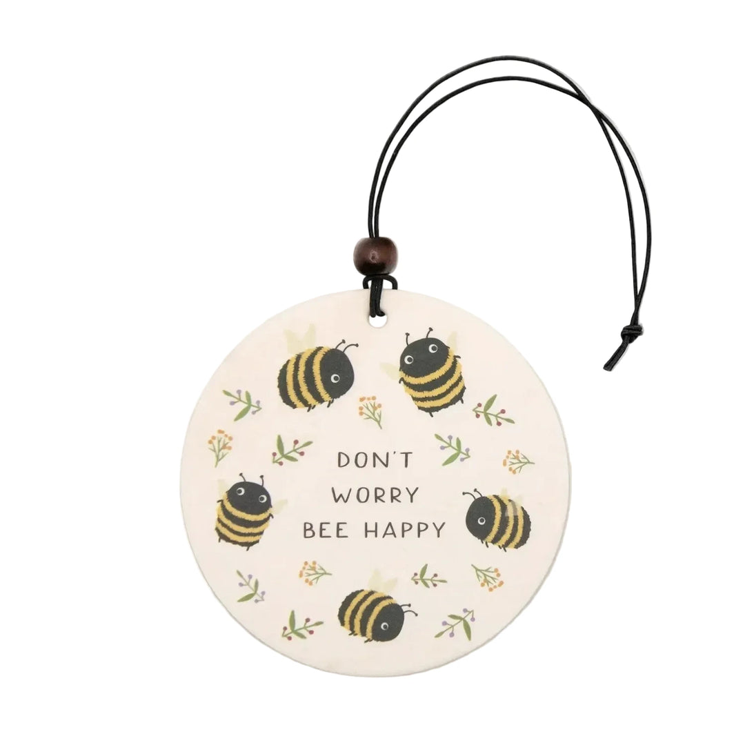 A round cream-colored car air freshener printed with five chubby cartoon bumblebees and small wildflower sprigs surrounding the text "Don't Worry Bee Happy," hanging from a black cord with a dark wooden bead accent. 