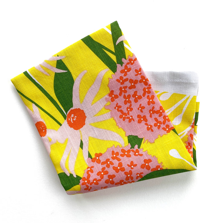 Folded tea towel showcasing close-up of the colorful floral print featuring pink daisies and orange alliums with green stems.