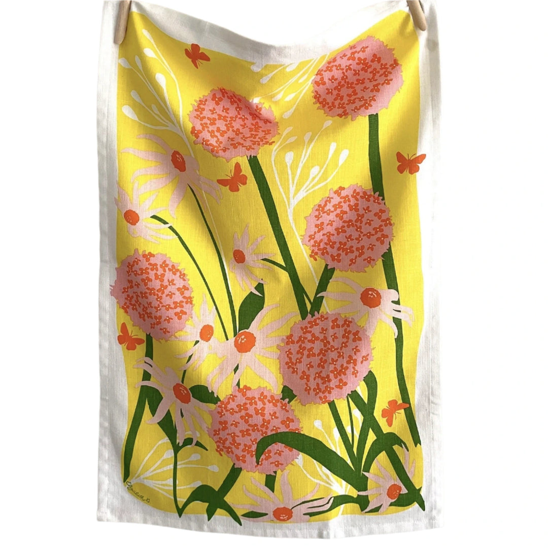 Tea towel hanging on a line, printed with a vibrant allium and daisy floral pattern in pink, orange, and green on a yellow background with a white border.
