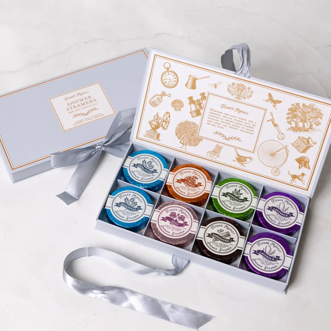 A set of eight aromatherapy shower steamers in a gift box with a satin ribbon. The steamers are in shades of blue, green, purple, and brown, each labeled with different essential oils.