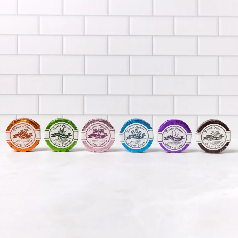 6 steamers (Lavender, Eucalyptus, Rose, Citrus, Peppermint, and Sandalwood scents) each in colorful foil wrapping with round white branded label displayed on a white ledge in front of a wall of white subway tiles.