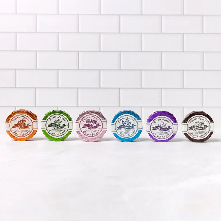 6 steamers (Lavender, Eucalyptus, Rose, Citrus, Peppermint, and Sandalwood scents) each in colorful foil wrapping with round white branded label displayed on a white ledge in front of a wall of white subway tiles.