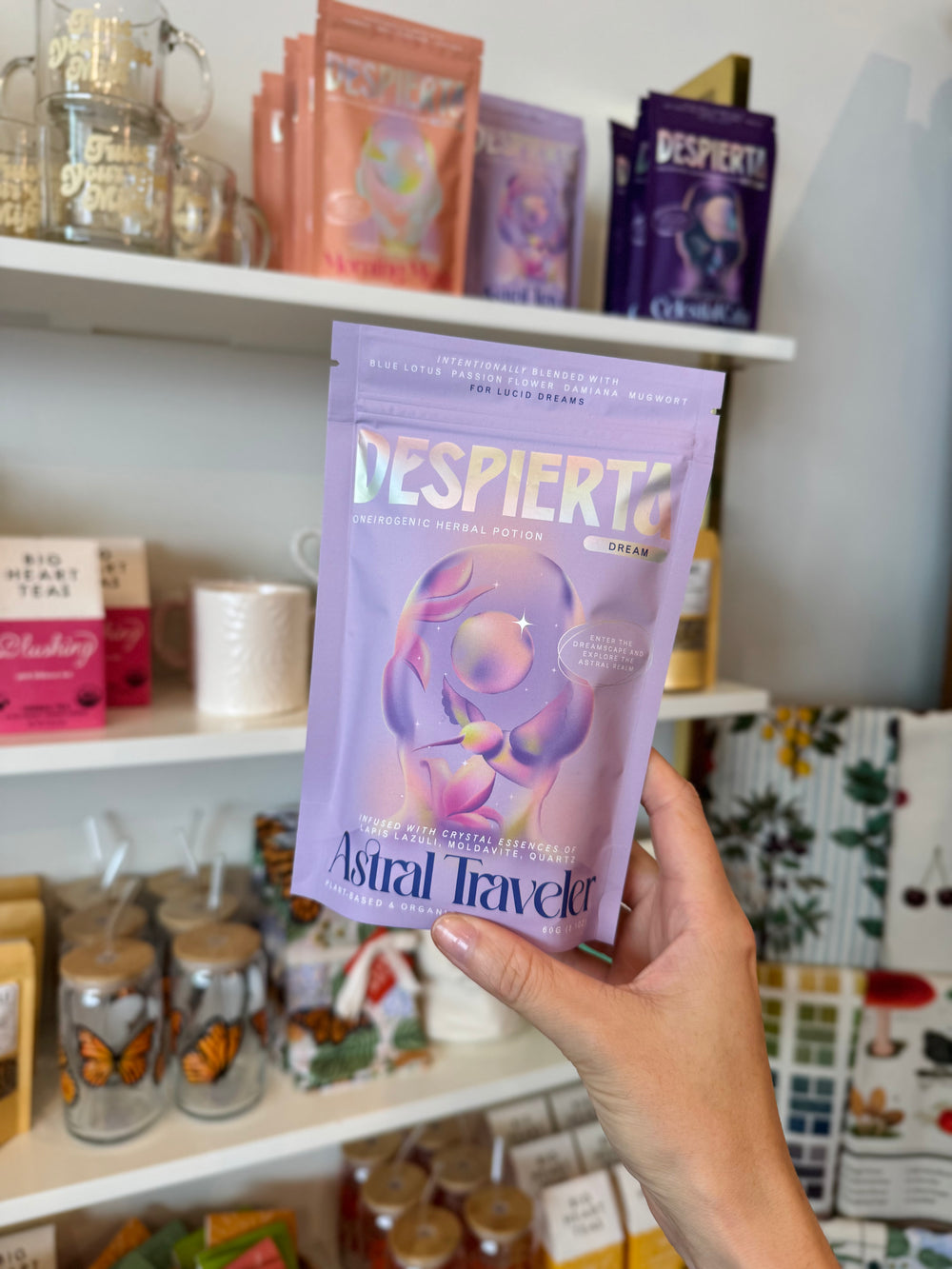 Hand holding a Despierta Astral Traveler package in front of a shelf with various products.