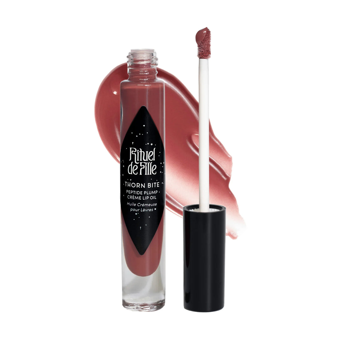 clear tube of rose shaded lip oil with a black branded label and lip with a swatch of the lip oil behind it. Brand: Rituel De Fille