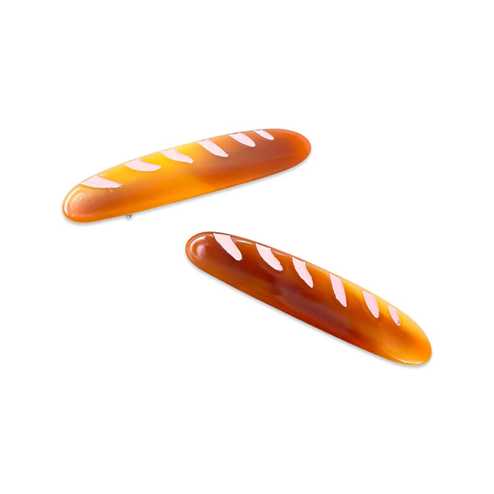 Two glossy, baguette-shaped alligator hair clips with a golden brown design and white diagonal marks, styled to resemble French bread.