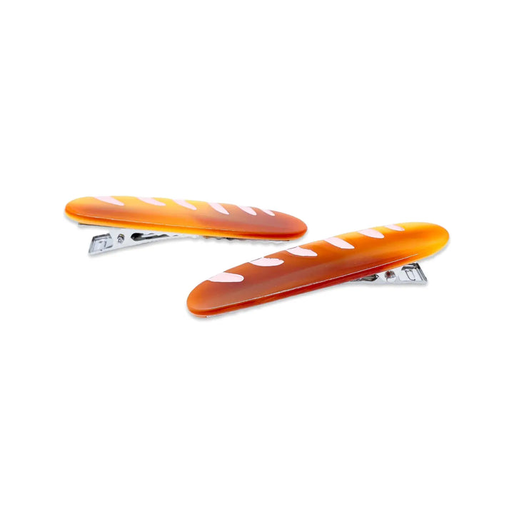Two glossy, baguette-shaped alligator hair clips with a golden brown design and white diagonal marks, styled to resemble French bread.