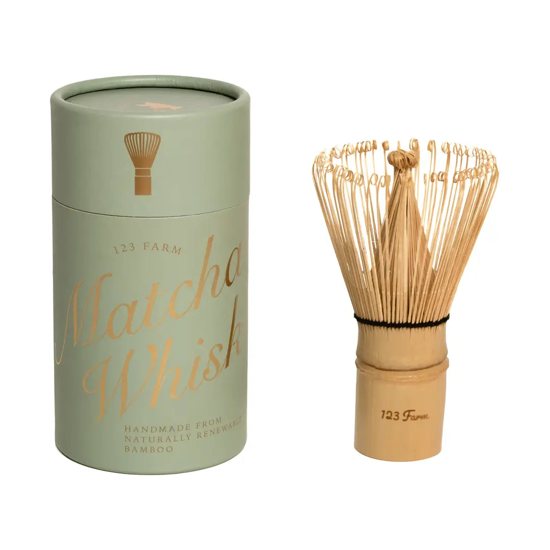 A handmade bamboo matcha whisk chasen by 123 Farm next to its sage green and gold cylindrical gift box.
