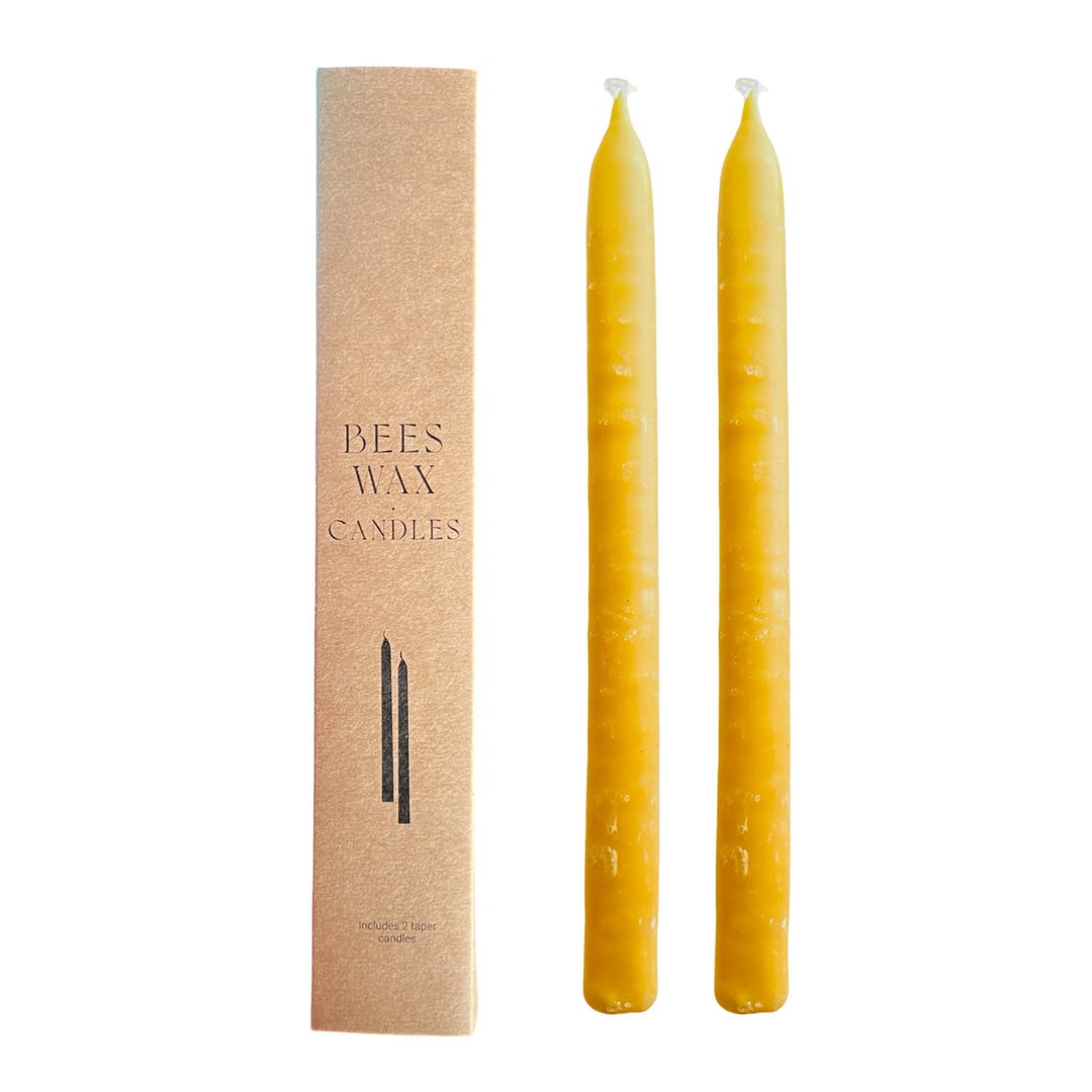A set of two yellow beeswax taper candles alongside their packaging, which is a simple rectangle with the product name 'Beeswax Taper Candles' printed on it.