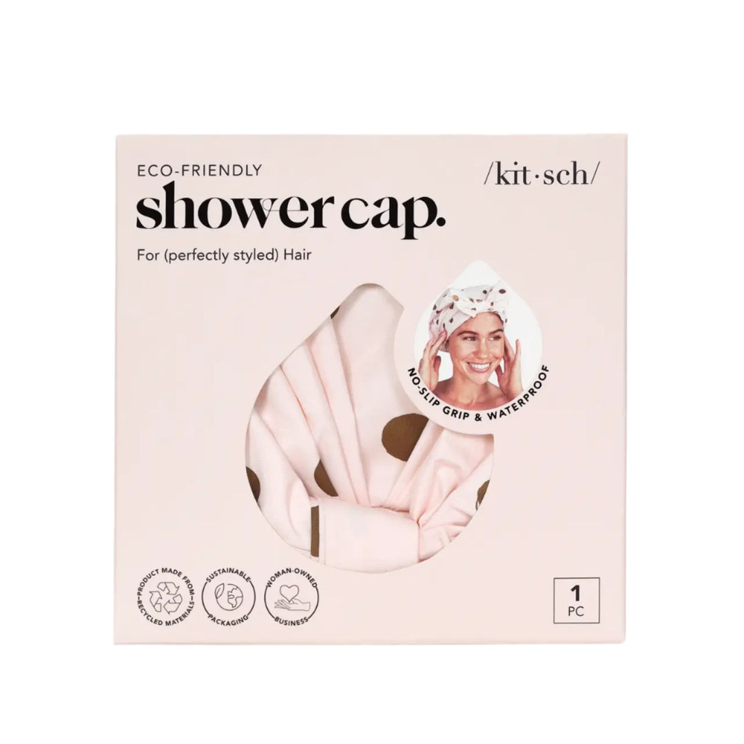 A Kitsch Luxury Shower Cap – Blush Dot in its eco-friendly packaging. The packaging is soft pink with a cutout window displaying the shower cap’s blush-pink fabric with gold polka dots. A small circular image in the top right corner shows a woman wearing the cap, highlighting its no-slip grip and waterproof design. Icons on the bottom indicate that the product is made from recycled materials, features sustainable packaging, and is from a woman-owned business.