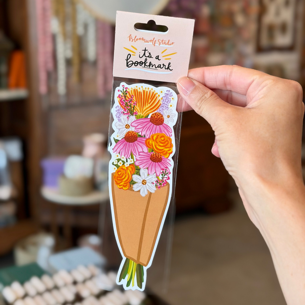 A tall vertical die cut bookmark illustrated as a wrapped paper bouquet of pink coneflowers, orange zinnias, white daisies, protea, lavender, and wax flowers with green stems.