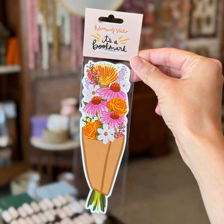 A tall vertical die cut bookmark illustrated as a wrapped paper bouquet of pink coneflowers, orange zinnias, white daisies, protea, lavender, and wax flowers with green stems.