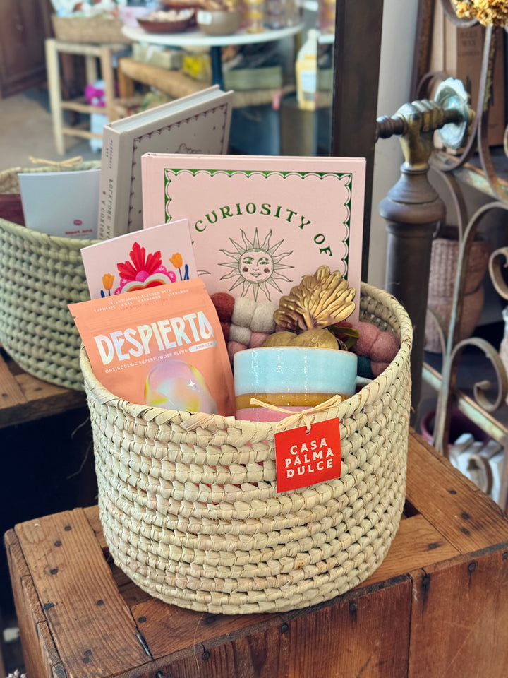 A handwoven palm leaf Casa Palma Dulce gift basket filled with a Despierta superpowder blend packet, a ceramic cup, a candle, a book, and decorative items, displayed on a wooden crate.