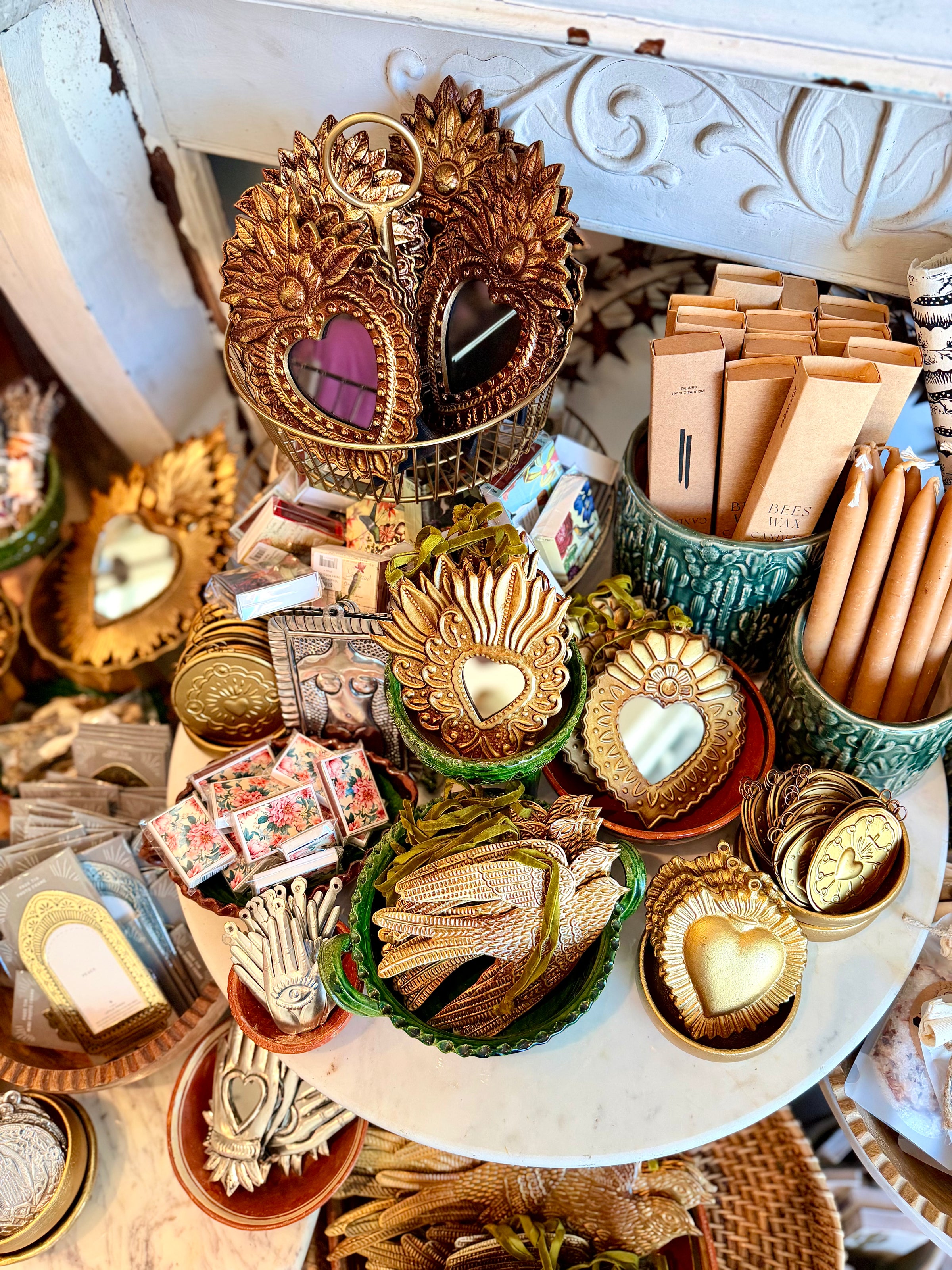 A styled tabletop display of decorative gold-toned heart-shaped mirrors, small heart-shaped ornaments, packaged accessories, and green ceramic holders filled with sticks or pencils, arranged in a boutique-like setting.