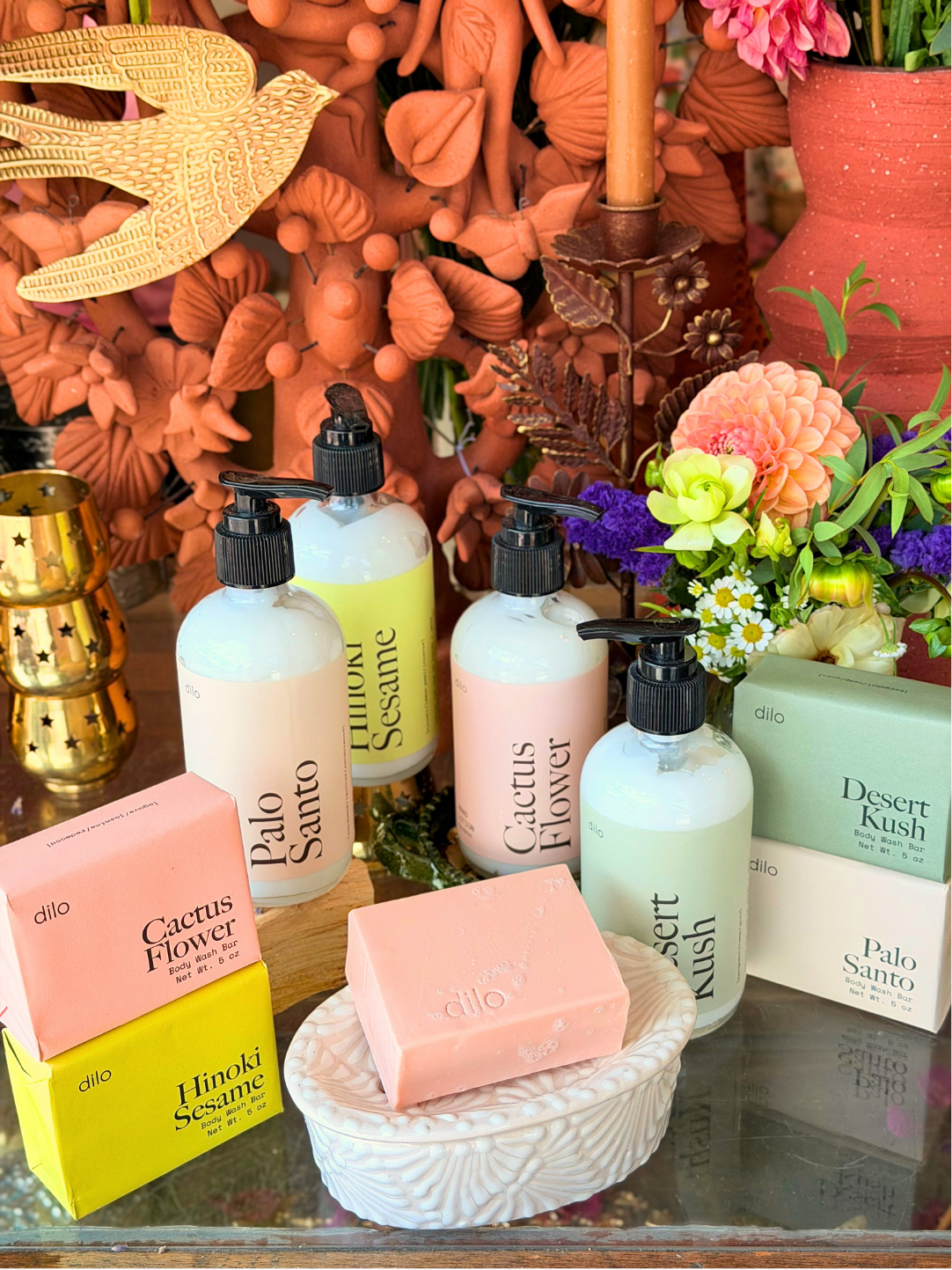 A styled product display of Dilo brand skincare and bath products, including pump bottles of Sesame, Cactus Flower, and Desert Kush, alongside a pink soap bar in a textured dish and matching product boxes, set against a warm terracotta decorative background.