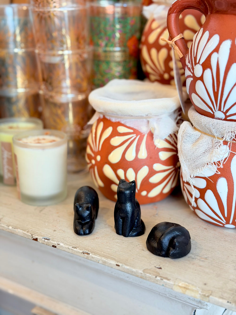 A styled tabletop display featuring a terracotta-colored ceramic pitcher and jars with white floral patterns, alongside three small black cat figurines in the foreground. 