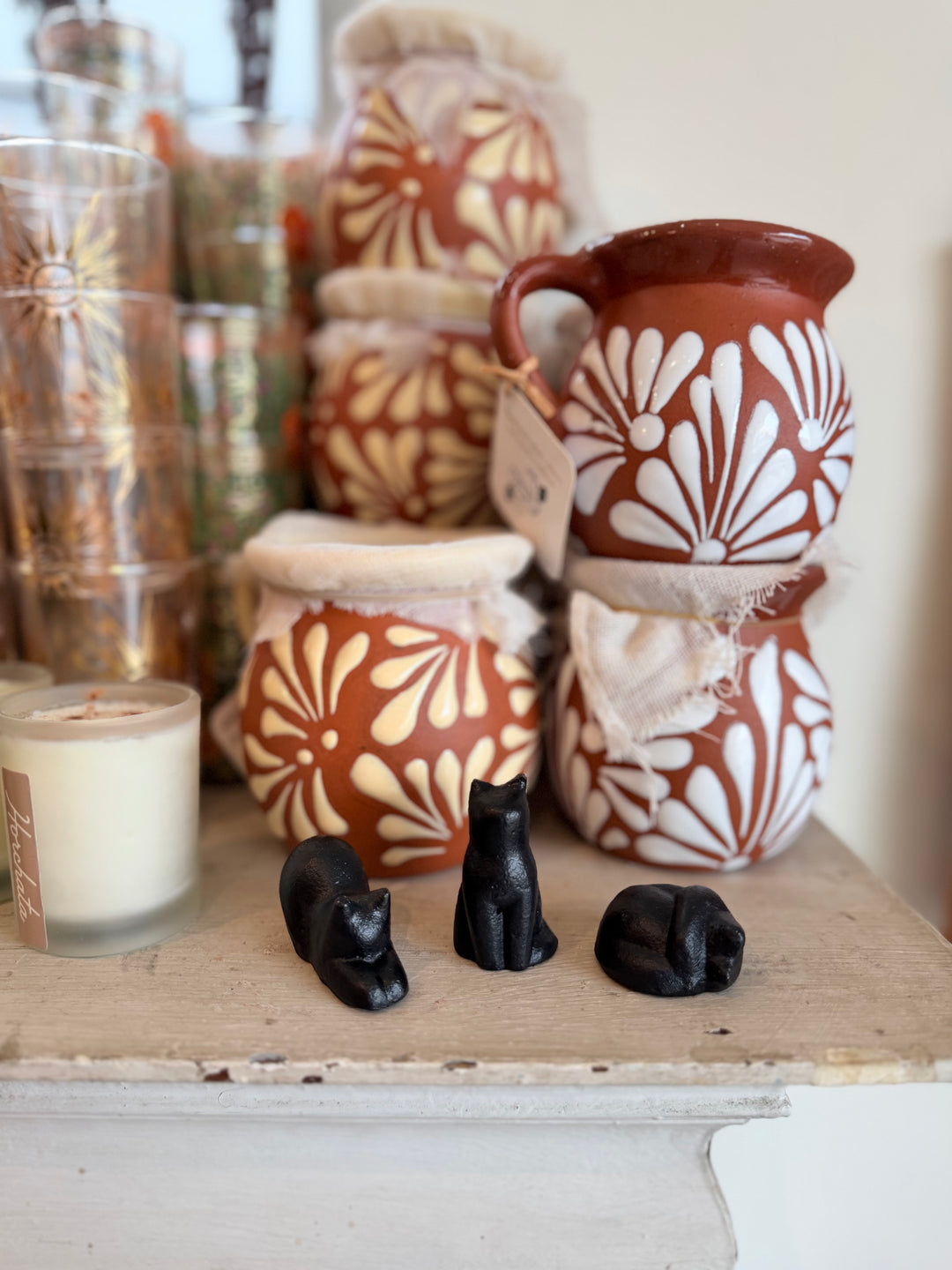 A styled tabletop display featuring a terracotta-colored ceramic pitcher and jars with white floral patterns, alongside three small black cat figurines in the foreground. 