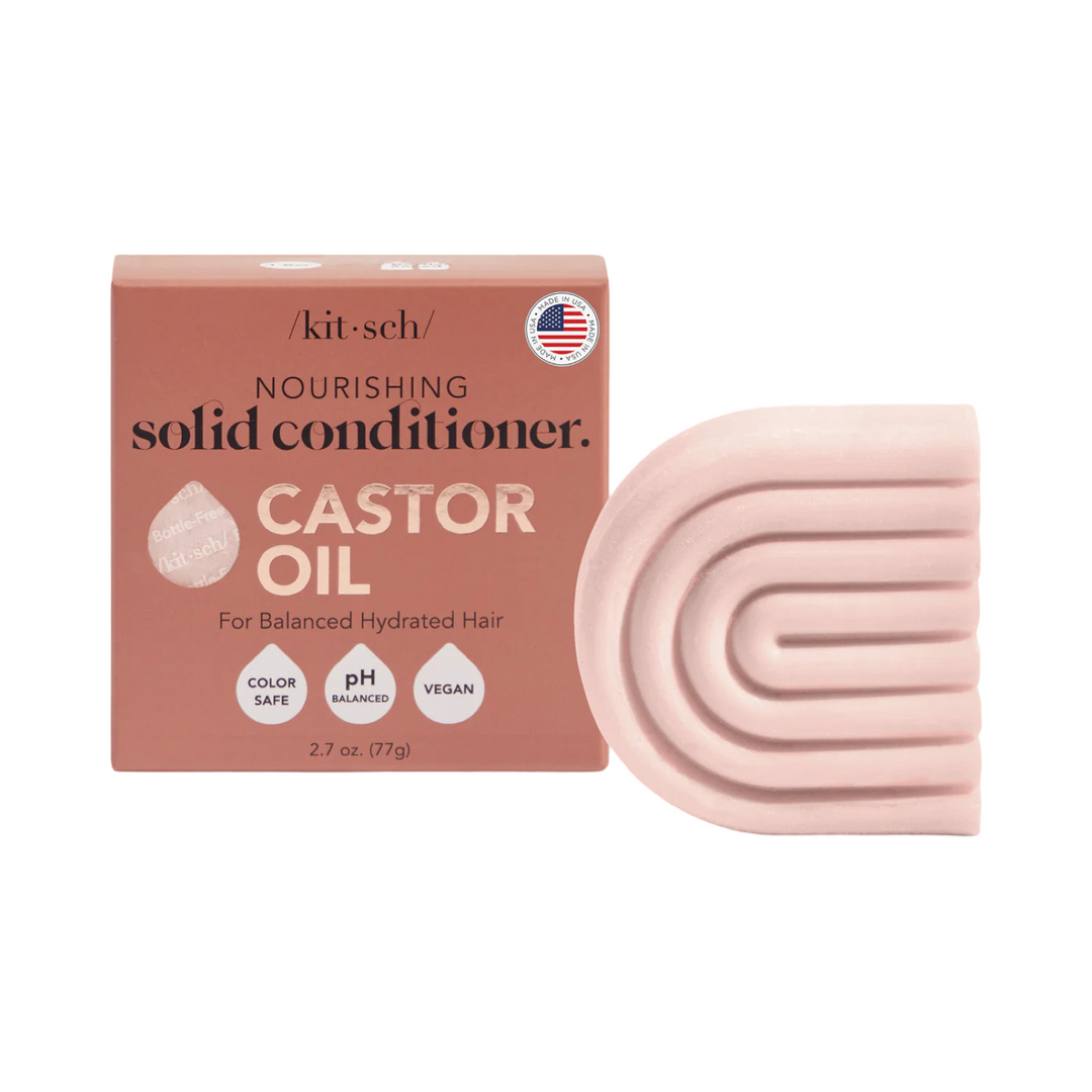 A Kitsch Castor Oil Nourishing Solid Conditioner Bar alongside its packaging, which is a soft peach color with gold lettering. The packaging highlights key features like color-safe, pH-balanced, and vegan, and includes a "Made in the USA" label. The conditioner bar itself has a curved, textured design.