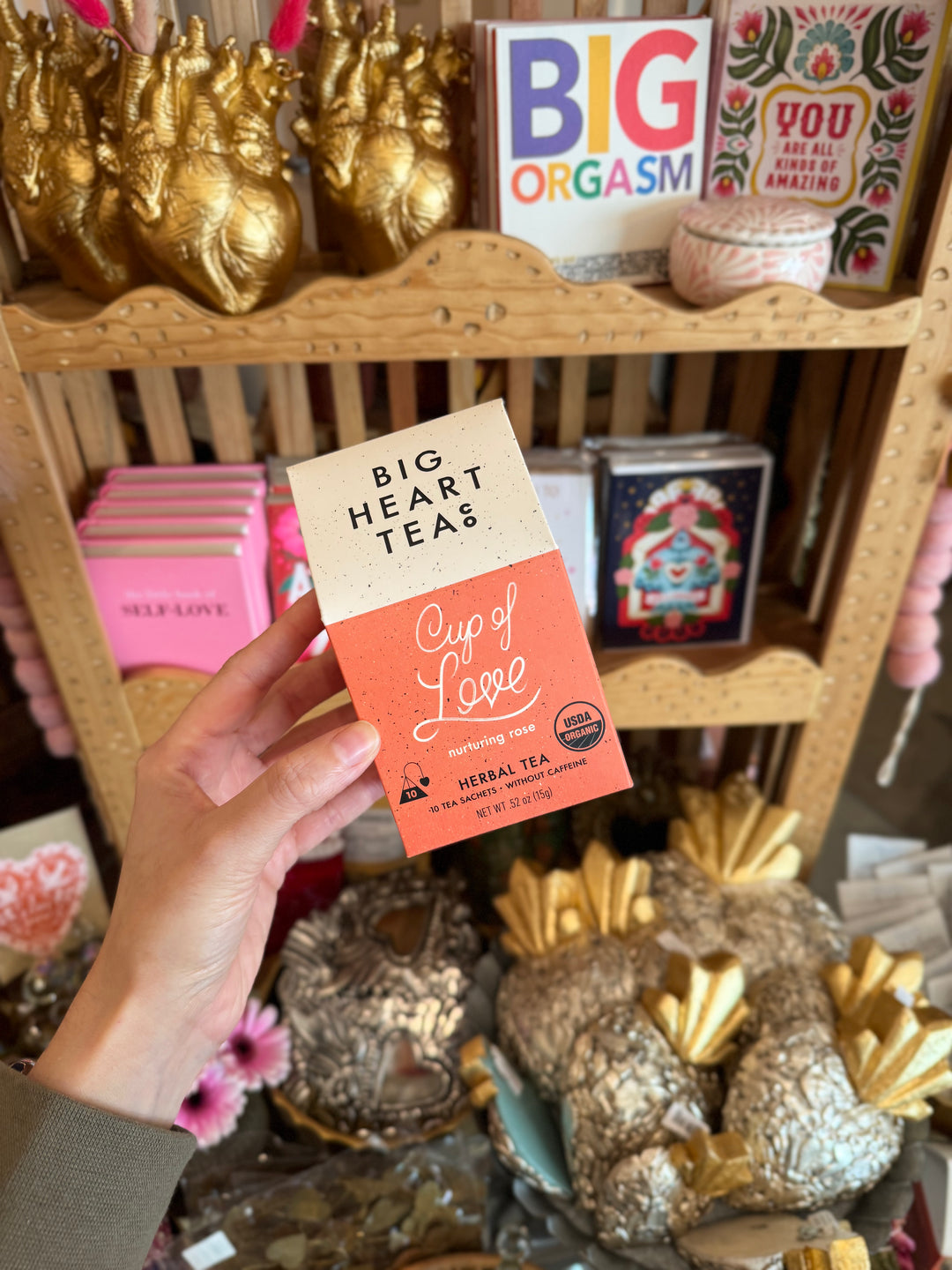 Person holding a 'Big Heart Teas Cup of Love' package in front of a shelf with decorative items.