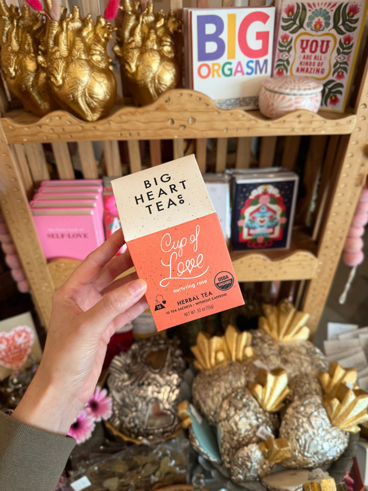 Person holding a 'Big Heart Teas Cup of Love' package in front of a shelf with decorative items.
