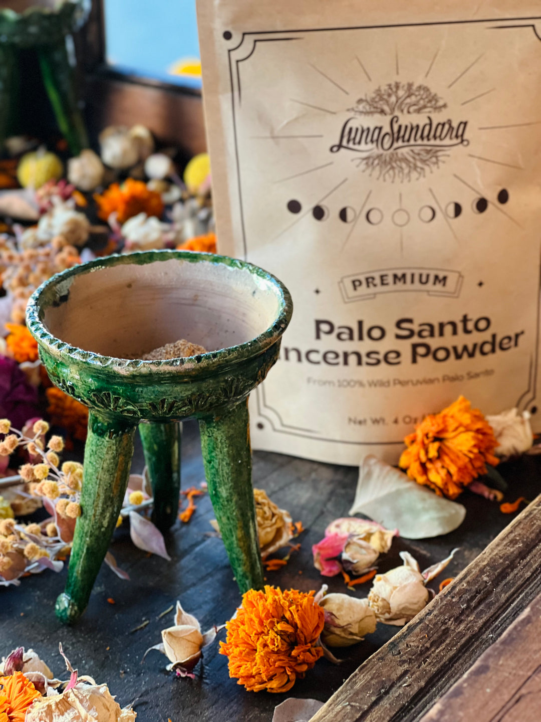 A bag of Luna Sundara Premium Palo Santo Incense Powder stands behind a small green ceramic copalero filled with incense powder, surrounded by dried flowers and botanicals on a dark wooden surface.