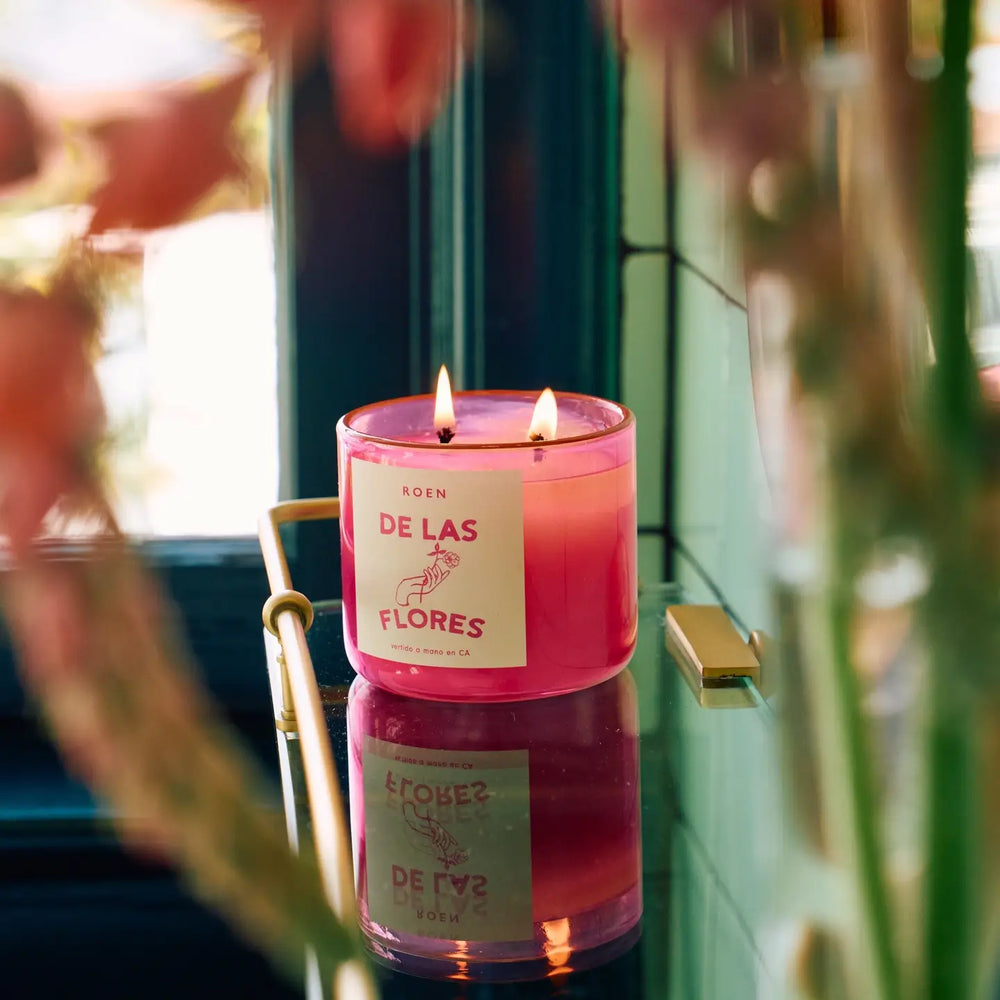 Photo of burning De Las Flores candle on a glass shelf in front of a green background.