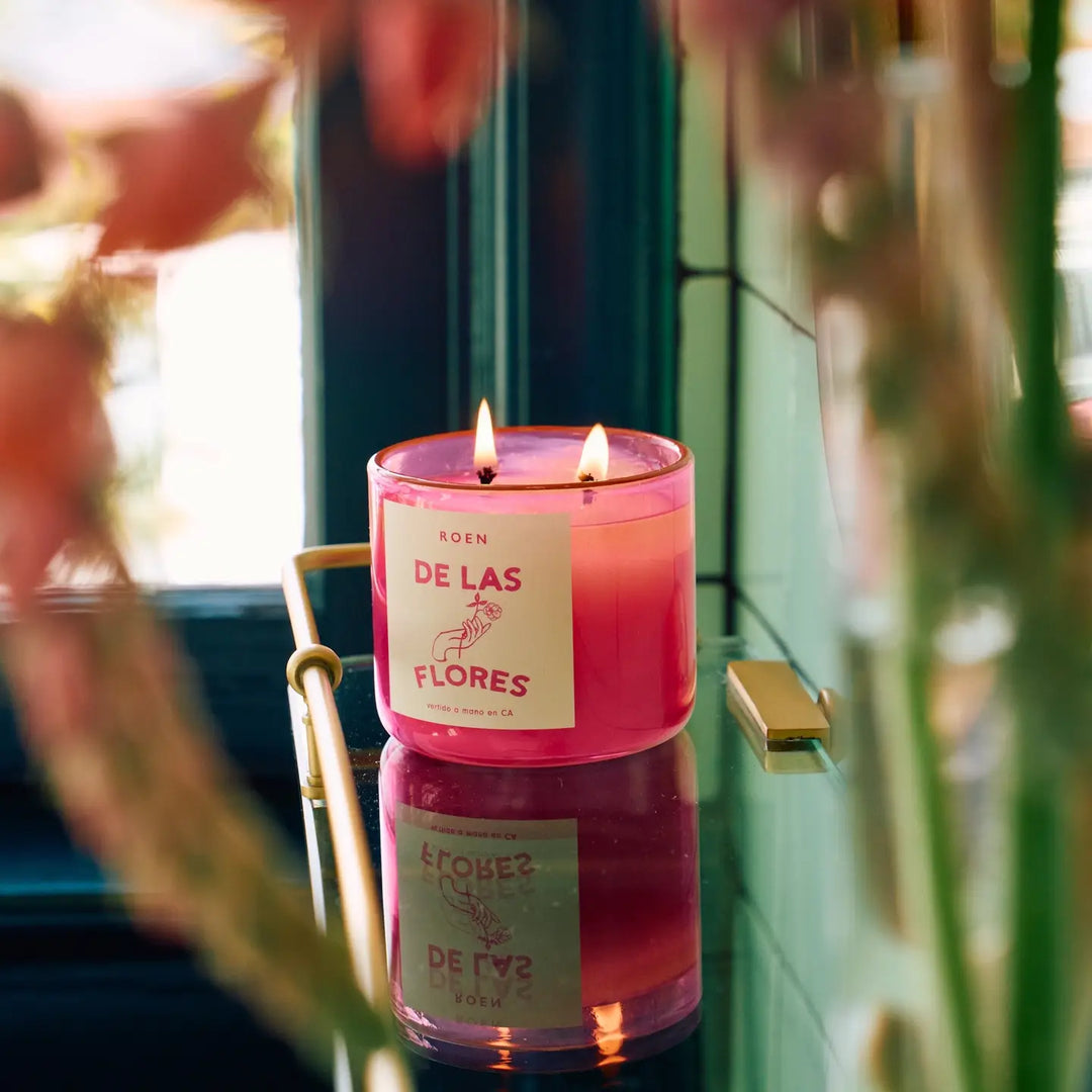 Photo of burning De Las Flores candle on a glass shelf in front of a green background.
