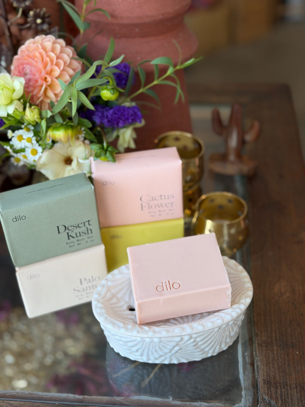 4 Dilo scented soap bars in pastel-colored boxes (Desert Kush, Cactus Flower, Palo Santo, Hinoki Sesame) arranged on a glass tray next to a white ceramic dish with a pink soap bar, set against a rustic background with flowers and brass candleholders.