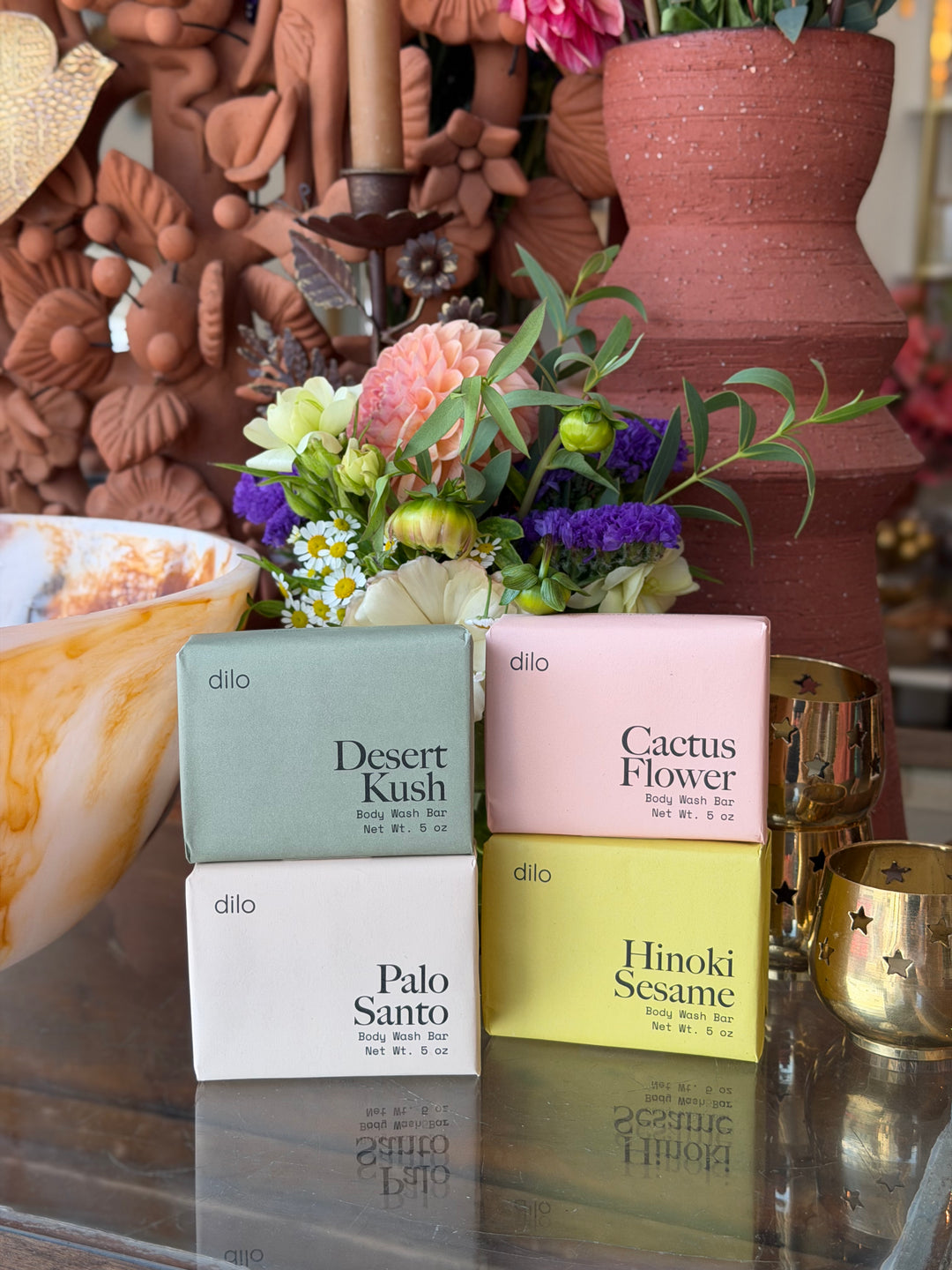 Four Dilo brand body soap boxes in different colors & scents arranged on a reflective glass table in front of a decorative floral arrangement and terracotta vases.