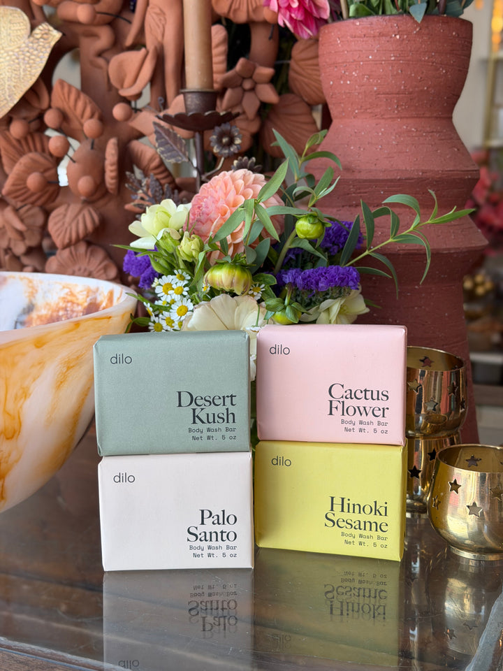Four Dilo brand body soap boxes in different colors & scents arranged on a reflective glass table in front of a decorative floral arrangement and terracotta vases.