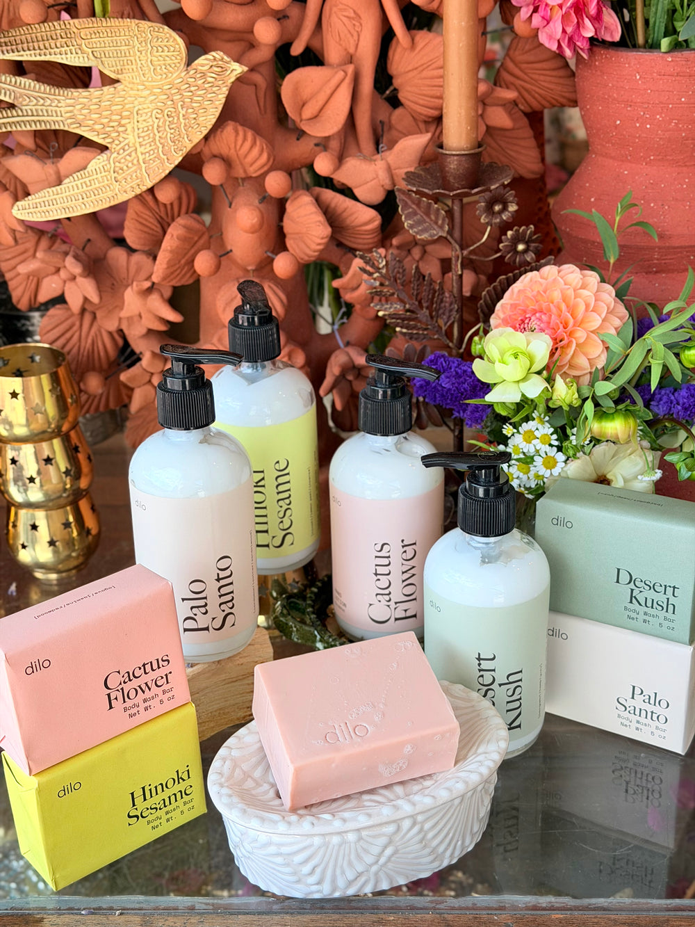 A styled product display of Dilo brand bathroom and body care items, including pump bottles labeled Palo Santo, Hinoki Sesame, Cactus Flower, and Desert Kush, alongside matching boxes and a pink soap bar in a white talavera soap dish.