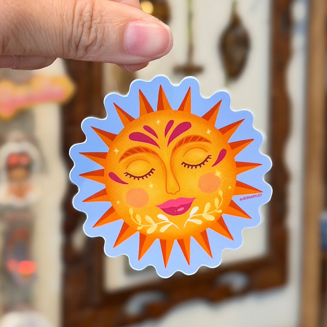 Hand holding a colorful sun-shaped sticker with a blurred background of art supplies.