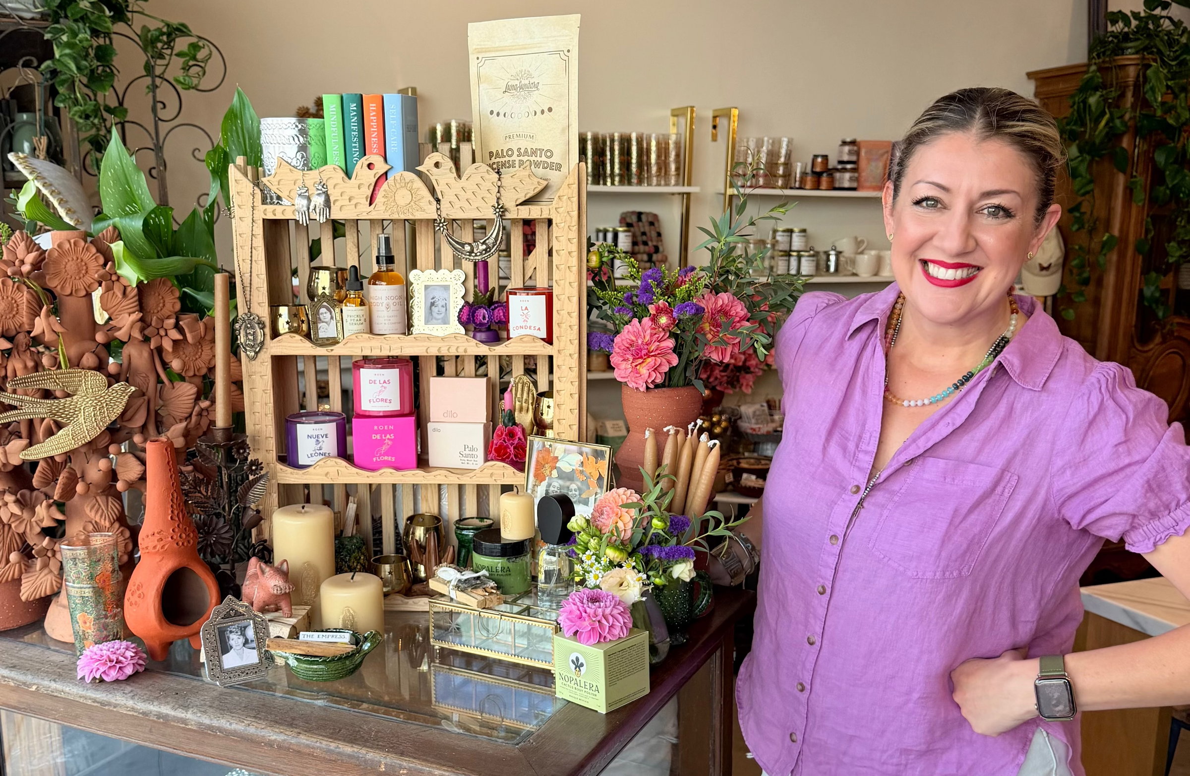 A woman smiling and posing next to a wooden display cabinet in a shop filled with candles, flowers, and decorative items. 