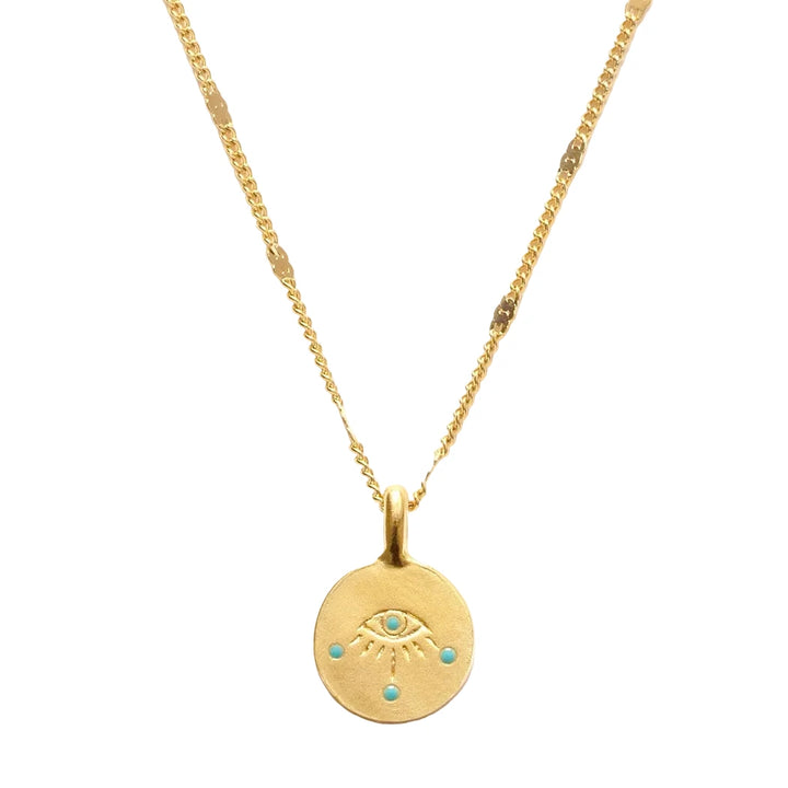 A close-up of a small round gold coin pendant featuring an engraved evil eye with turquoise enamel dot accents, hung on a delicate mixed gold chain with flat bar and curb link details.