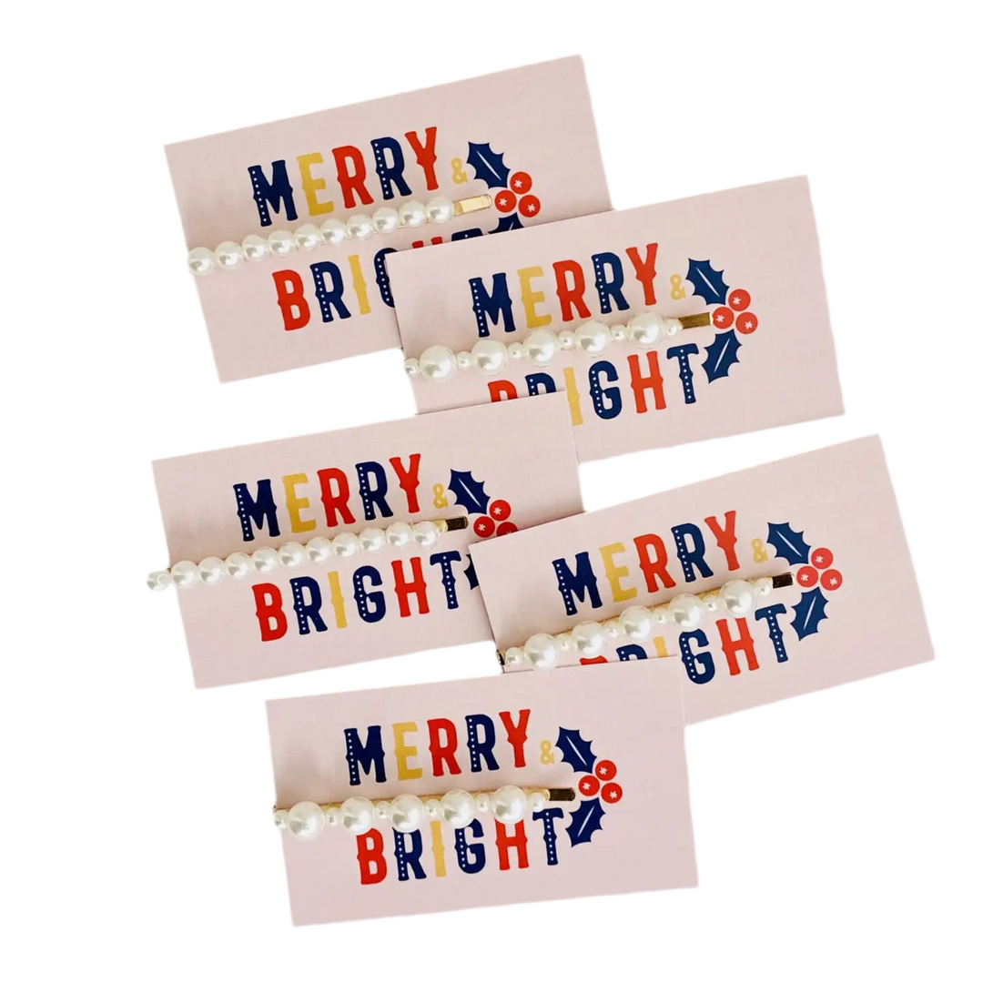 A set of pearl-embellished hairpins displayed on festive pink cards with the words “Merry & Bright” in colorful lettering and holly berry accents.