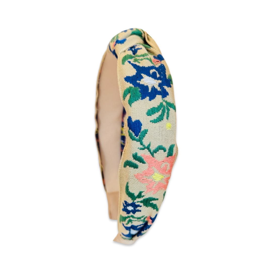 Alternate side view of a wide knotted fabric headband with a natural linen-textured base, covered in colorful floral embroidery featuring cobalt blue, green, peach, and yellow botanical motifs. The headband has a center knot detail and smooth cream inner lining.