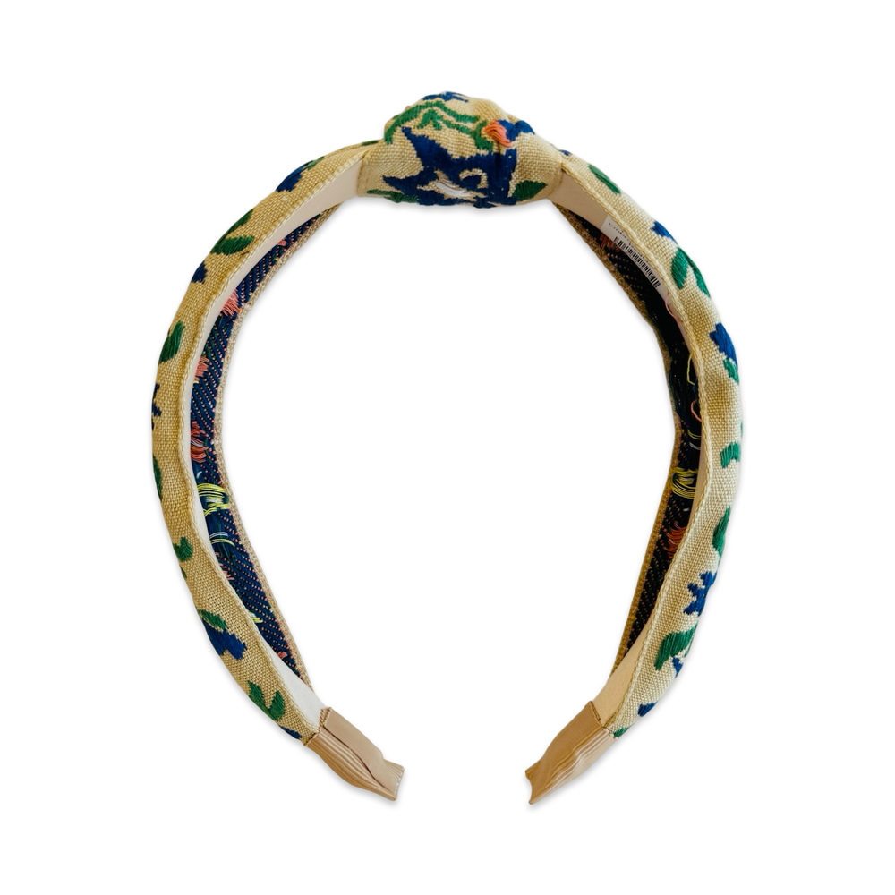 Front view of a wide knotted fabric headband with a natural linen-textured base, covered in colorful floral embroidery featuring cobalt blue, green, peach, and yellow botanical motifs. The headband has a center knot detail and smooth cream inner lining.